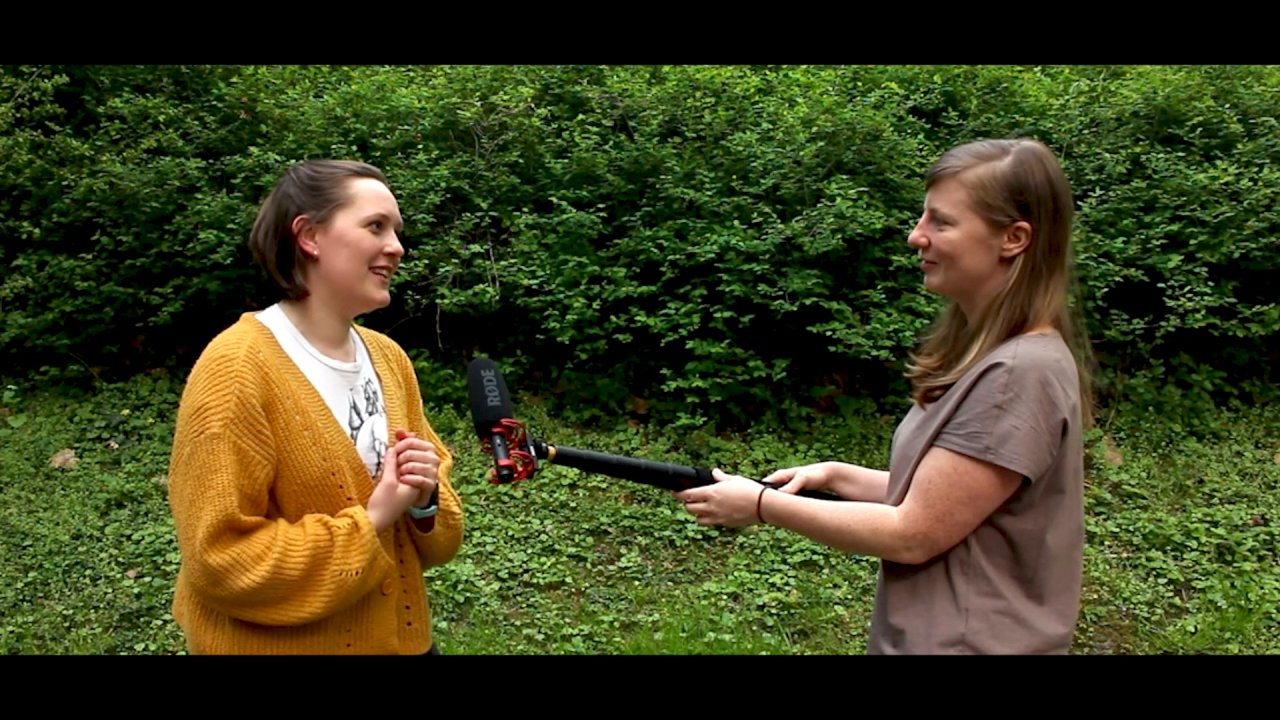Lisa interviewt Alina im Park mit einem Mikrofon in der Hand. Im Hintergrund sind Büsche.